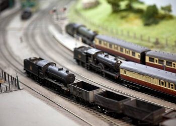 "Model railway layout. A freight train with a model steam locomotive passes two passenger trains at a station. Shallow depth of field, focus on the foreground."