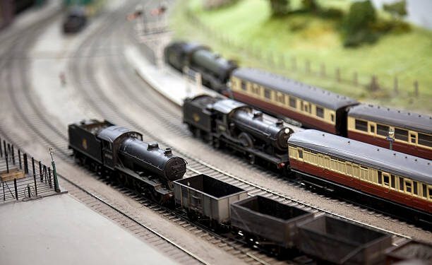 "Model railway layout. A freight train with a model steam locomotive passes two passenger trains at a station. Shallow depth of field, focus on the foreground."